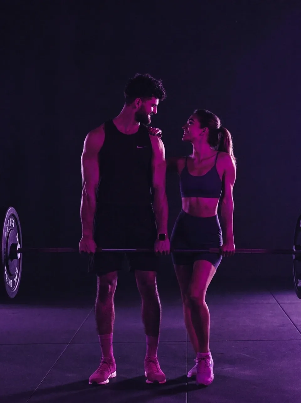 Casal apoiado numa barra de musculação, sob luz roxa